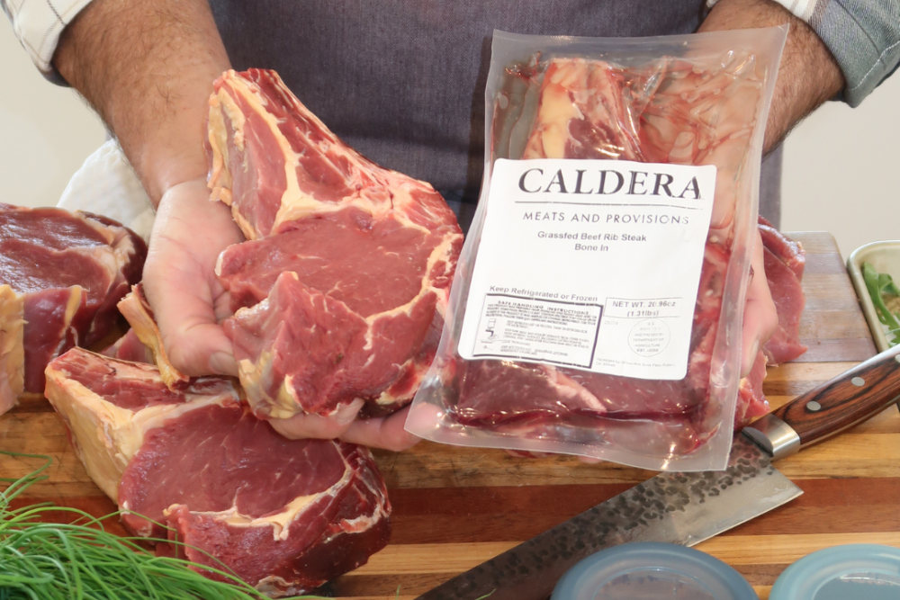 Person holding a package of raw meat labeled 'Caldera' with various meats and ingredients on a cutting board.