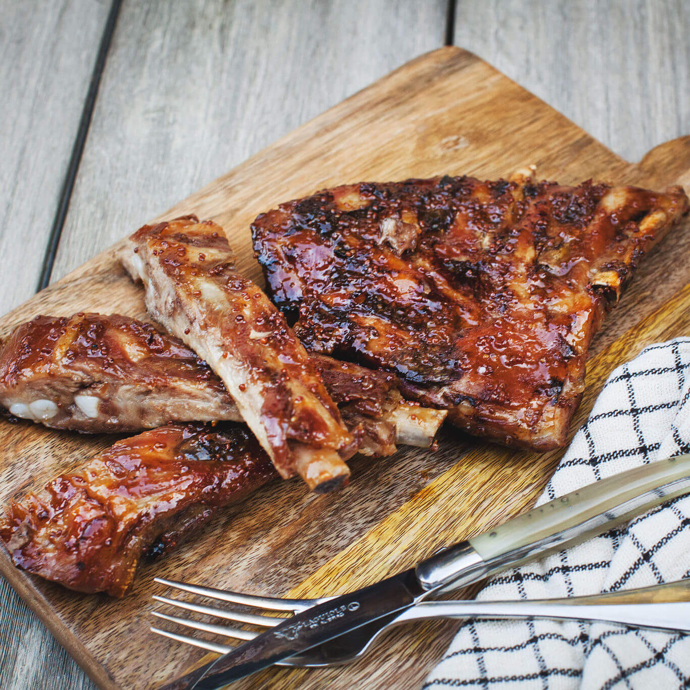 Barbecue ribs on a wooden cutting board with utensils on a rustic wooden surface